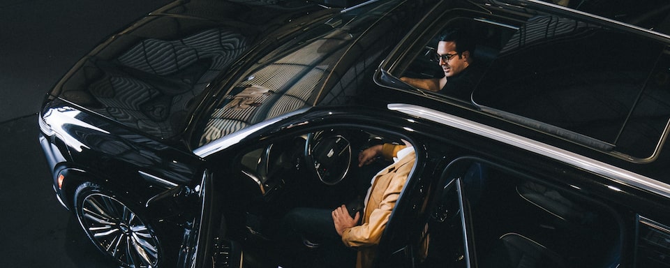 Dois homens embarcados em um SUV Cadillac vistos de cima; um deles, inclusive, tem o rosto revelado pelo sofisticado teto solar panorâmico, que se abre qual um sorriso dos deuses da engenharia automobilística.