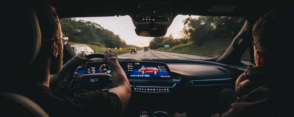 Visão interna do painel de um veículo Cadillac em movimento na estrada. Além do motorista e da passageira, vemos todo o painel do carro, a central multimídia, o painel de controle, o volante, e o espelho retrovisor.