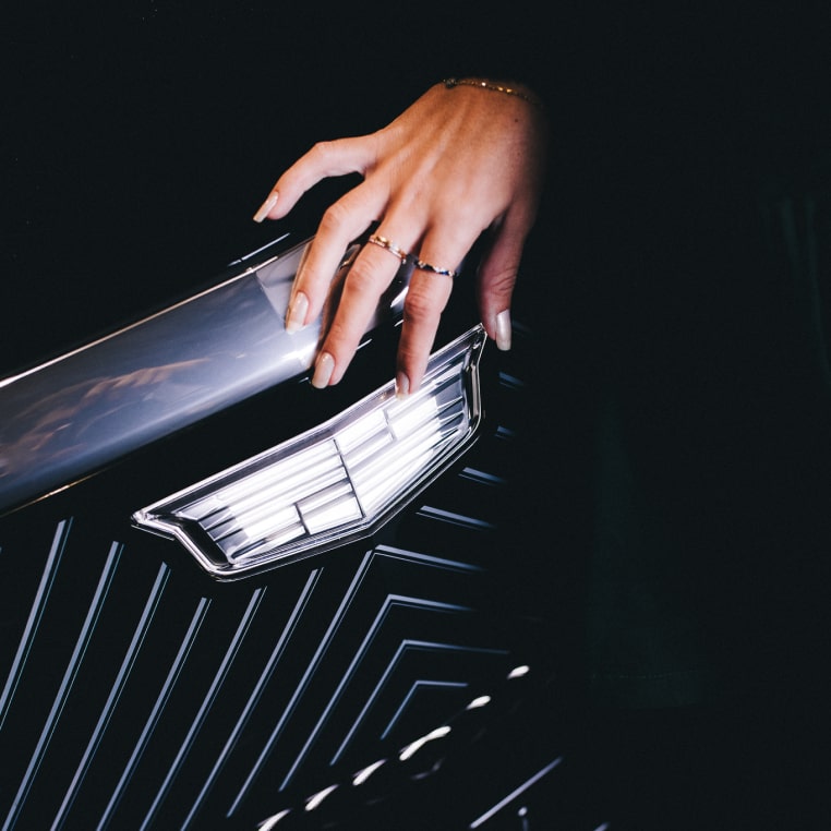 Detalhe do emblema Cadillac, aplicado na grade frontal de um dos veículos da marca, sendo tocado por mãos femininas com unhas prateadas e brilhantes.