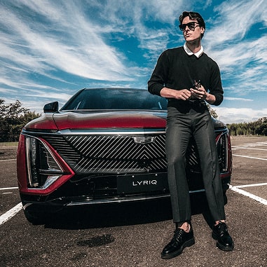 Um modelo está posando em frente ao Lyriq. Ele usa camisa, calças e sapatos pretos, contrastando com o vermelho do carro. 