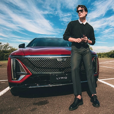 Um modelo está posando em frente ao Lyriq. Ele usa camisa, calças e sapatos pretos, contrastando com o vermelho do carro. 