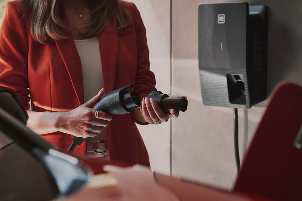 Mulher, elegantemente trajada com um blazer vermelho, em vias de conectar seu Cadillac 100% elétrico a um wall box.