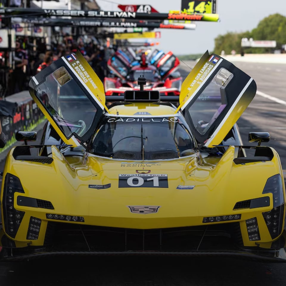 Frente de um carro de corrida Cadillac amarelo posicionado à beira da pista, com diversos outros em fileira logo atrás.