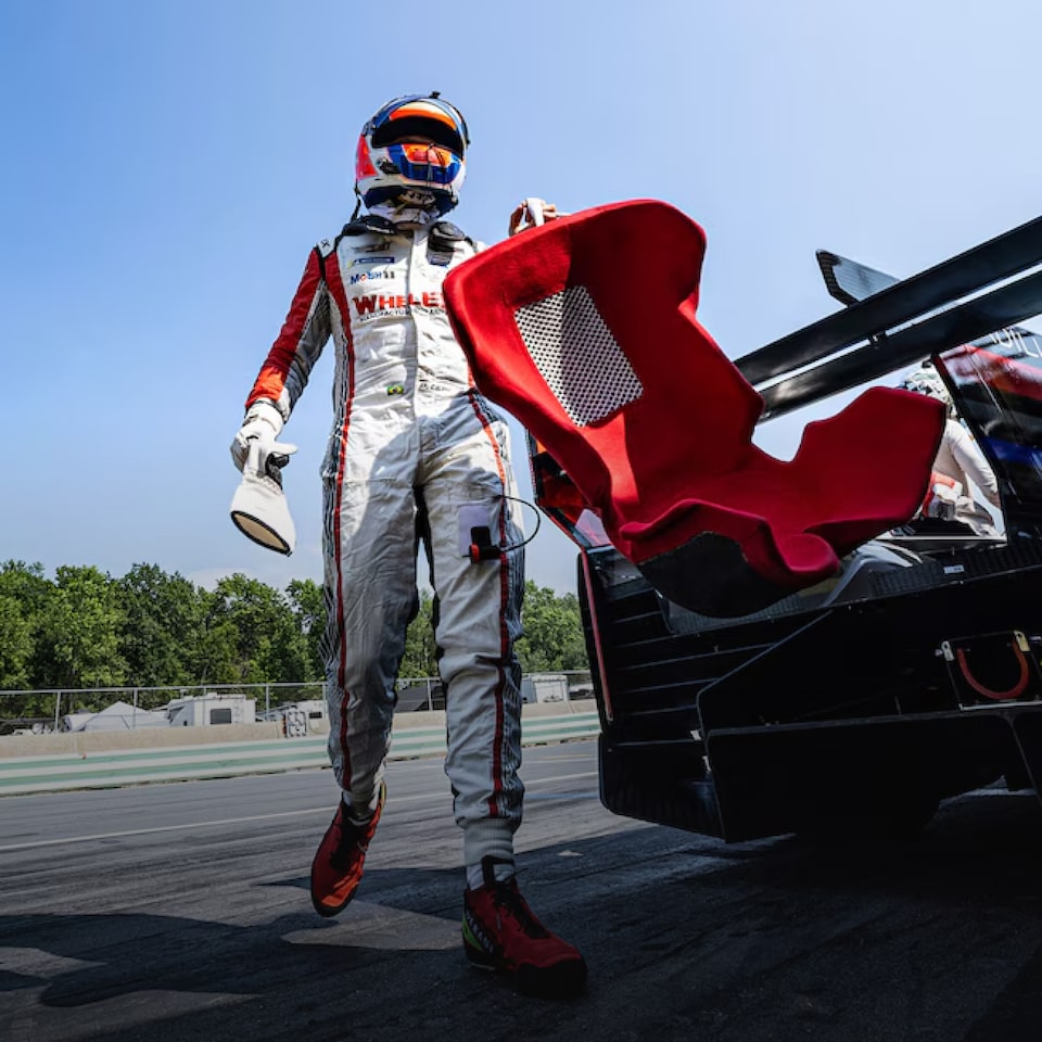 Pessoa em traje de corrida caminhando ao lado de um carro esportivo, pode-se ver a pista ao fundo. 