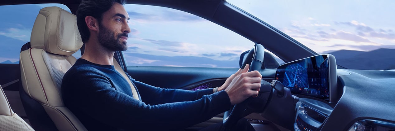 Motorista conduzindo um veículo Cadillac com interior premium, painel digital integrado, volante multifuncional e vista para a paisagem ao entardecer através das janelas.