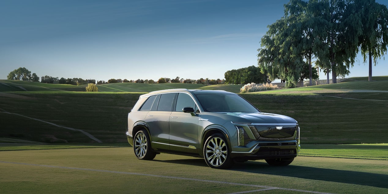 SUV elétrico Cadillac VISTIQ na cor cinza estacionado em uma área gramada, com árvores e paisagem ao fundo, destacando o design sofisticado, a grade frontal e as rodas de liga leve.
