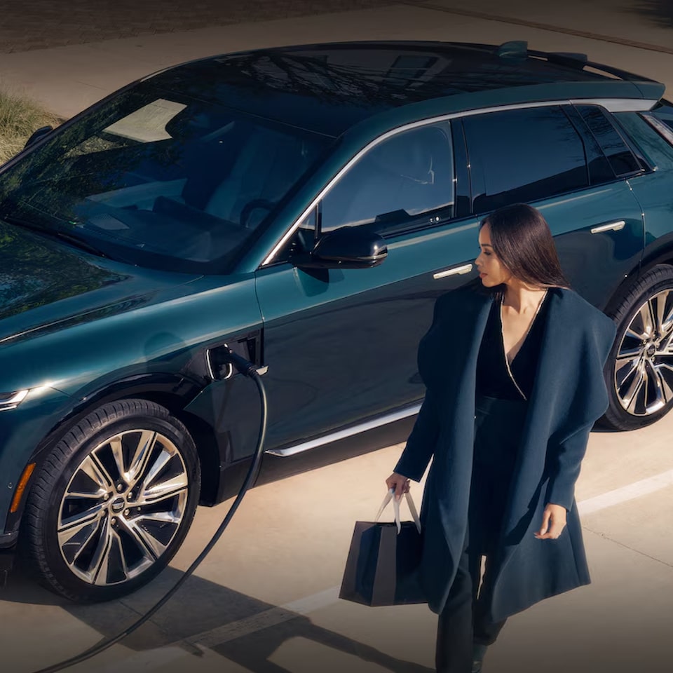 Imagem feita de cima de um Cadillac Lyriq sendo recarregado enquanto uma mulher de terno e sacolas nas mãos caminha ao seu lado.