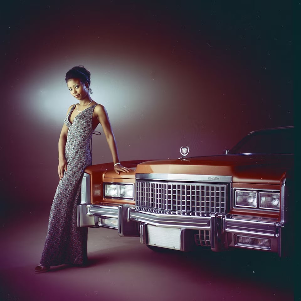 Mulher com um vestido prateado com a mão sob o capô do clássico Cadillac DeVille na cor vermelha.