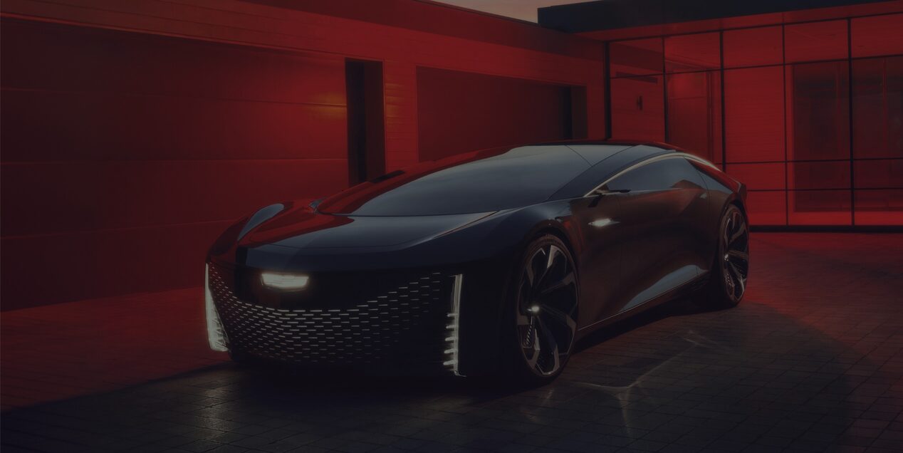 Imagem predominantemente escura, adornada por luzes e tons de vermelho ao fundo. No primeiro plano, um veículo Cadillac conceito às escuras, com destaque para as luzes full-LED dos faróis de condução diurna e do logo. No centro, o texto: "O extraordinário, à altura de Cadillac". No rodapé central, o aviso: "Os veículos aqui descritos não fazem parte do portfólio Brasil e não há previsão de chegada ao País".