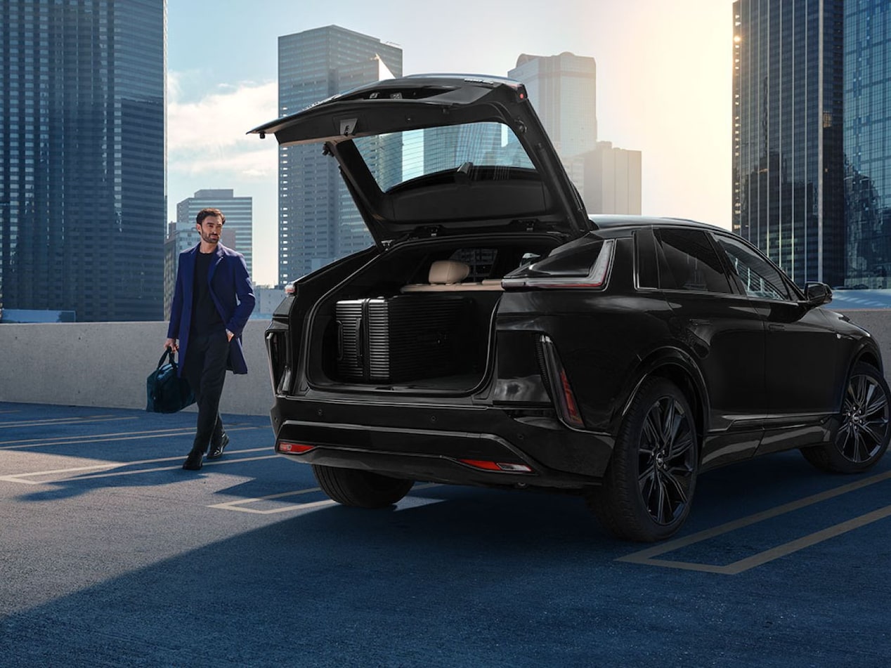 SUV Cadillac na cor preta estacionado em um ambiente urbano com o porta-malas aberto, enquanto uma pessoa caminha ao lado do veículo entre edifícios modernos.