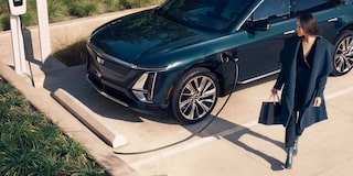 Cadillac LYRIQ na cor verde escuro conectado a um ponto de recarga para veículo elétrico enquanto uma mulher caminha ao lado do automóvel em um ambiente externo.