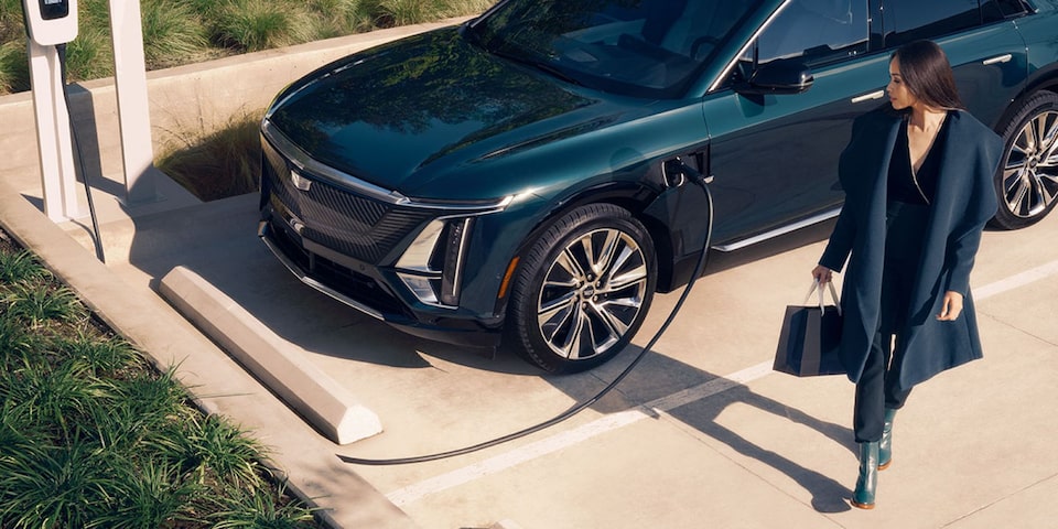 Cadillac LYRIQ na cor verde escuro conectado a um ponto de recarga para veículo elétrico enquanto uma mulher caminha ao lado do automóvel em um ambiente externo.