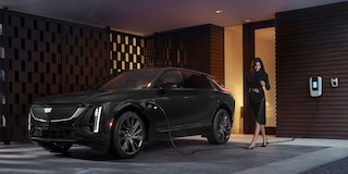 Cadillac LYRIQ na cor preta conectado a um carregador residencial enquanto uma mulher caminha ao lado do veículo em frente a uma residência.