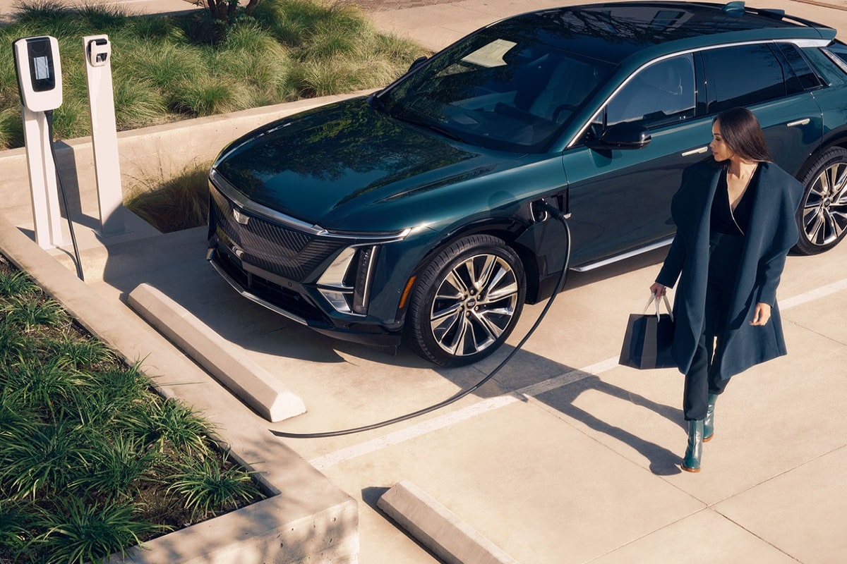 Cadillac LYRIQ na cor verde conectado a um ponto de recarga para veículo elétrico enquanto uma mulher caminha ao lado do veículo em um ambiente externo.