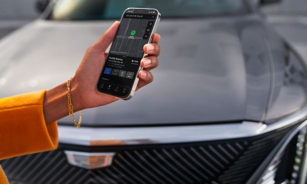 Pessoa utilizando o aplicativo myCadillac no smartphone em frente a um veículo Cadillac para acompanhar informações do veículo, status da bateria, localização, carregamento e autonomia.