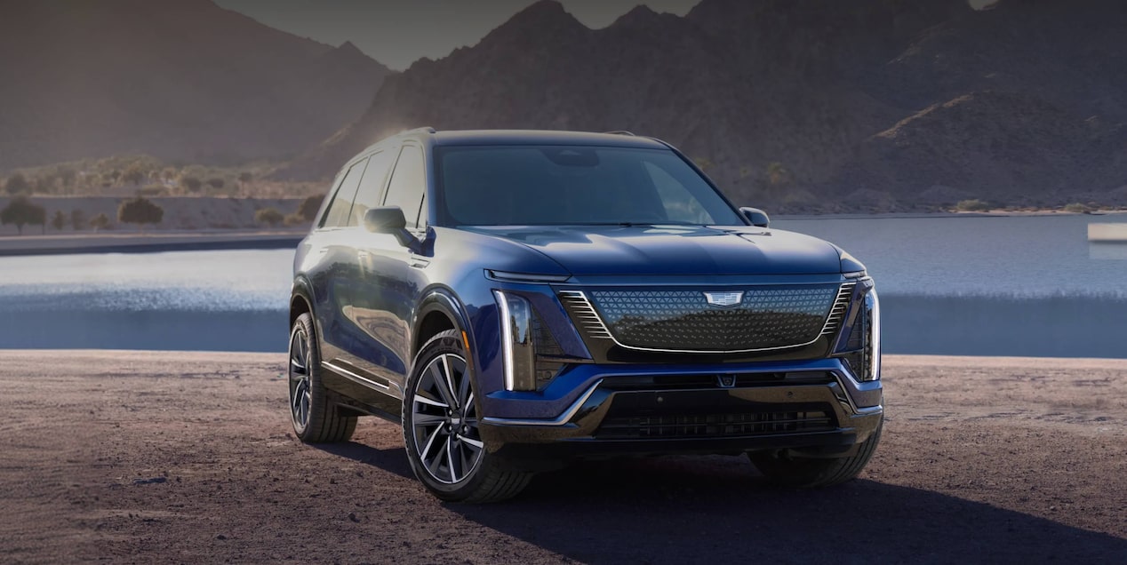 Cadillac Vistiq em ângulo 3/4 de frente, em imagem ambientada. O veículo aparece estacionado no pier, de costas para o mar e as belas montanhas verdejantes.