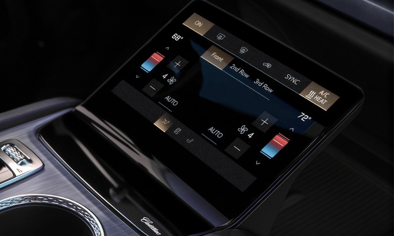 Console central de veículo Cadillac com tela de controles digitais, seletor giratório, porta-copos e acabamento premium em tons claros e escuros.