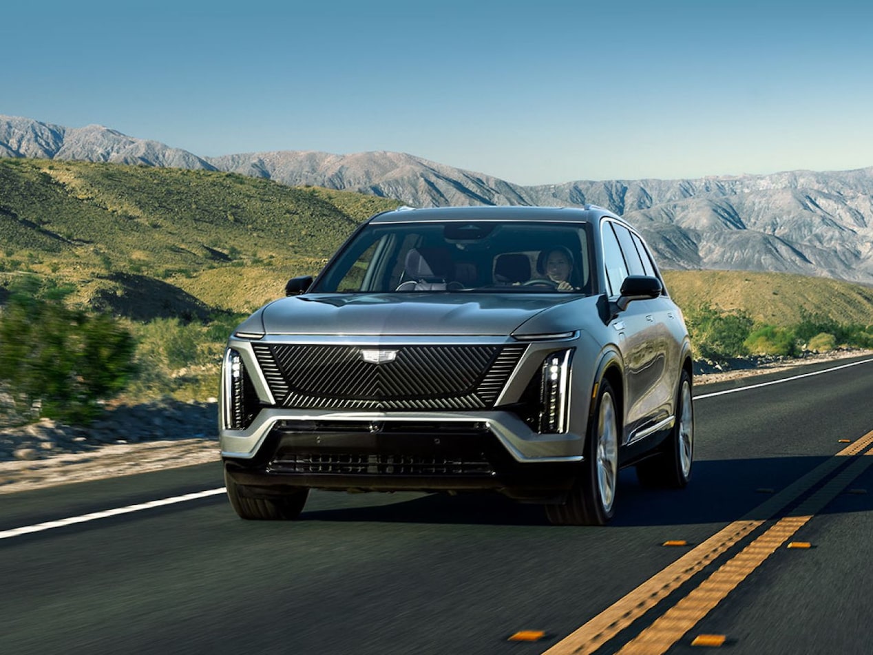 Carro elétrico Cadillac VISTIQ 2027 na cor cinza em movimento por uma estrada, exibido em visão frontal, com montanhas e vegetação ao fundo, destacando o design moderno e a iluminação em LED.
