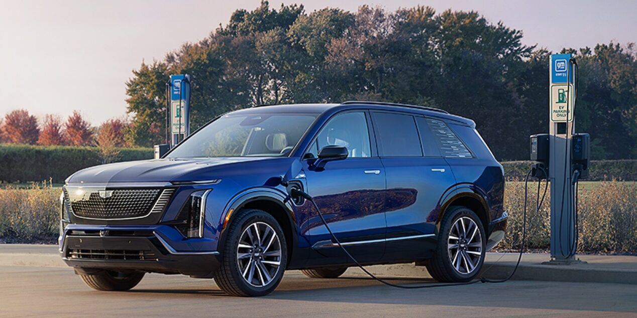 Alt: carro elétrico Cadillac na cor azul estacionado entre duas estações de carregamento para veículos elétricos, destacando o design moderno, as rodas de liga leve e a infraestrutura de recarga.