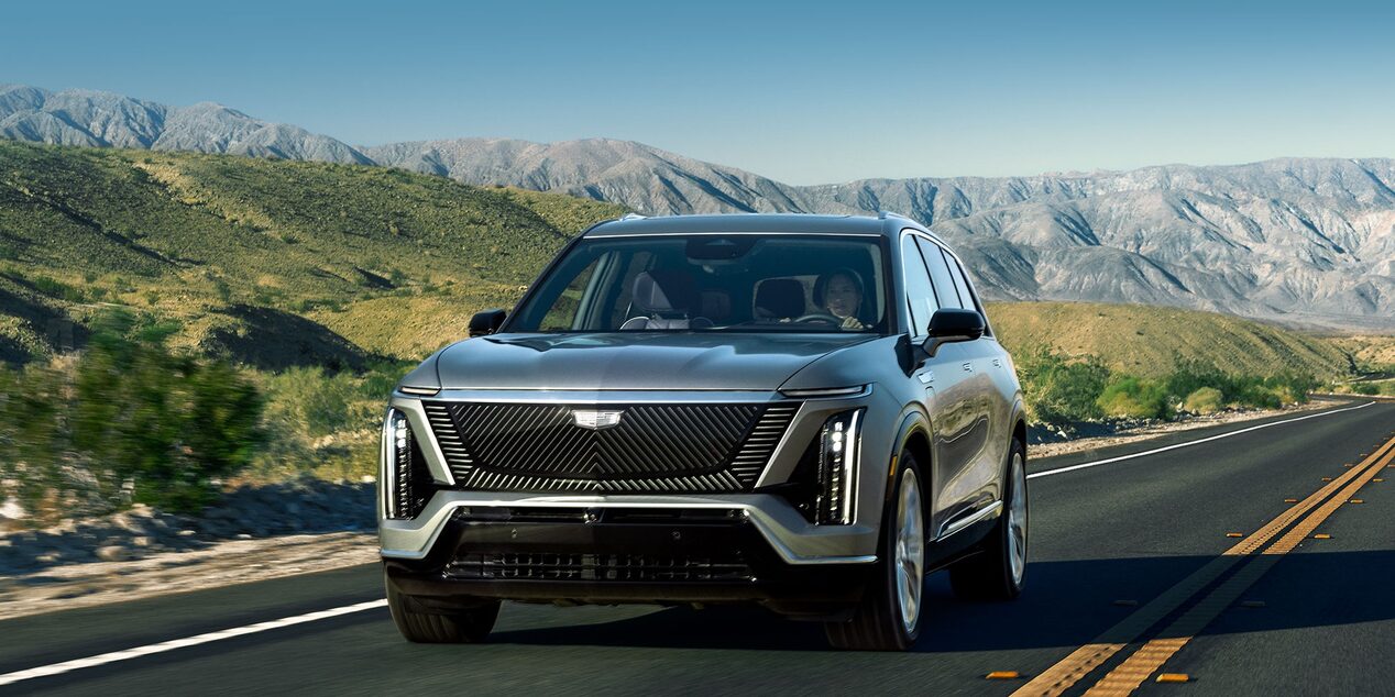 SUV elétrico Cadillac na cor cinza em movimento por uma estrada, exibido em visão frontal, com montanhas e vegetação ao fundo, destacando o design moderno e a iluminação em LED.