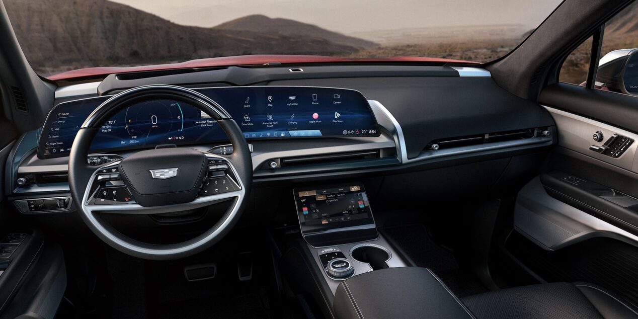 Interior de veículo Cadillac com painel digital integrado, volante multifuncional, central multimídia, console central e acabamento premium em tons escuros, com vista para uma paisagem montanhosa ao fundo.