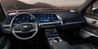 SUV elétrico Cadillac na cor cinza em movimento por uma estrada, exibido em visão frontal, com montanhas e vegetação ao fundo, destacando o design moderno e a iluminação em LED.