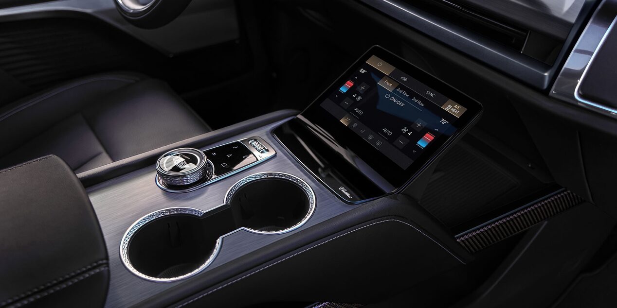 Console central de veículo Cadillac com porta-copos, seletor de controle, tela multimídia integrada e acabamento premium em tons escuros.