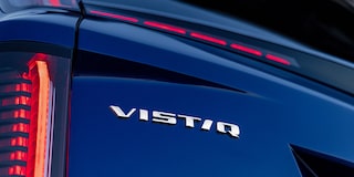Interior de veículo Cadillac com bancos em couro branco, painel digital integrado, volante multifuncional, console central e acabamento premium, com vista para a paisagem através do para-brisa.