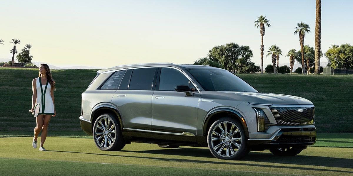 Carro Cadillac na cor cinza estacionado em uma área gramada, com uma mulher caminhando ao lado do veículo e árvores ao fundo, destacando o design sofisticado e as rodas de liga leve.