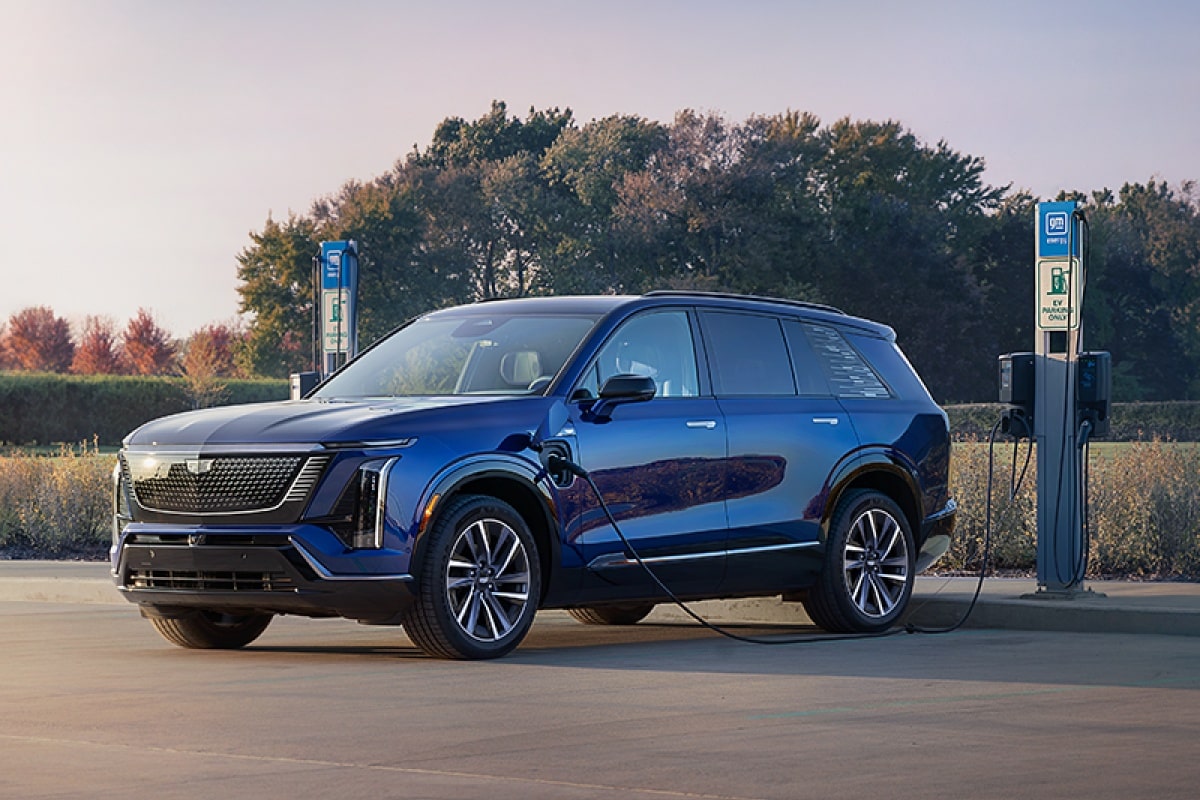 Carro elétrico Cadillac na cor azul estacionado entre duas estações de carregamento para veículos elétricos em uma área externa, com árvores e vegetação ao fundo, destacando o design moderno e a infraestrutura de recarga.